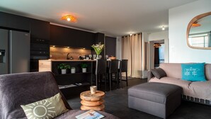 Un soggiorno moderno con un divano grigio, un pouf e un tavolino da caffè con una rivista. Si distingue un angolo cottura con mobili scuri e un piano di lavoro in legno. Sullo sfondo, una zona pranzo con tavolo e sedie.