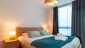 Un letto rifatto con cura, una coperta color petrolio, un comodino in legno con una lampada e un moderno lampadario.
