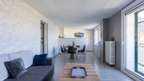 Een moderne woonkamer met een bank, eettafel en stoelen. Een radiator is rechts zichtbaar, en een raam biedt uitzicht op de omgeving.