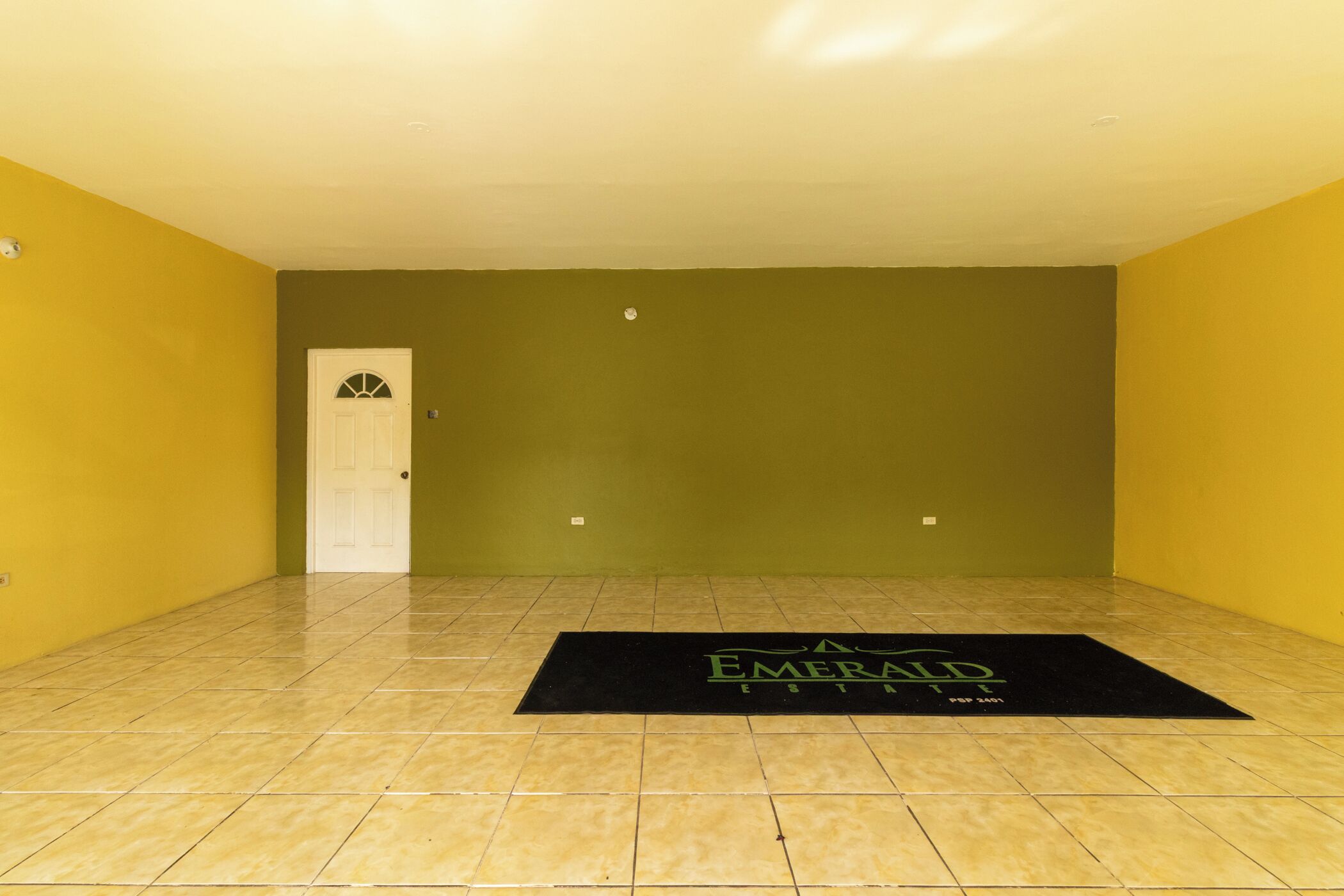 Een kamer met gele wanden, een witte deur en een zwarte mat met de tekst 'Emerald'.