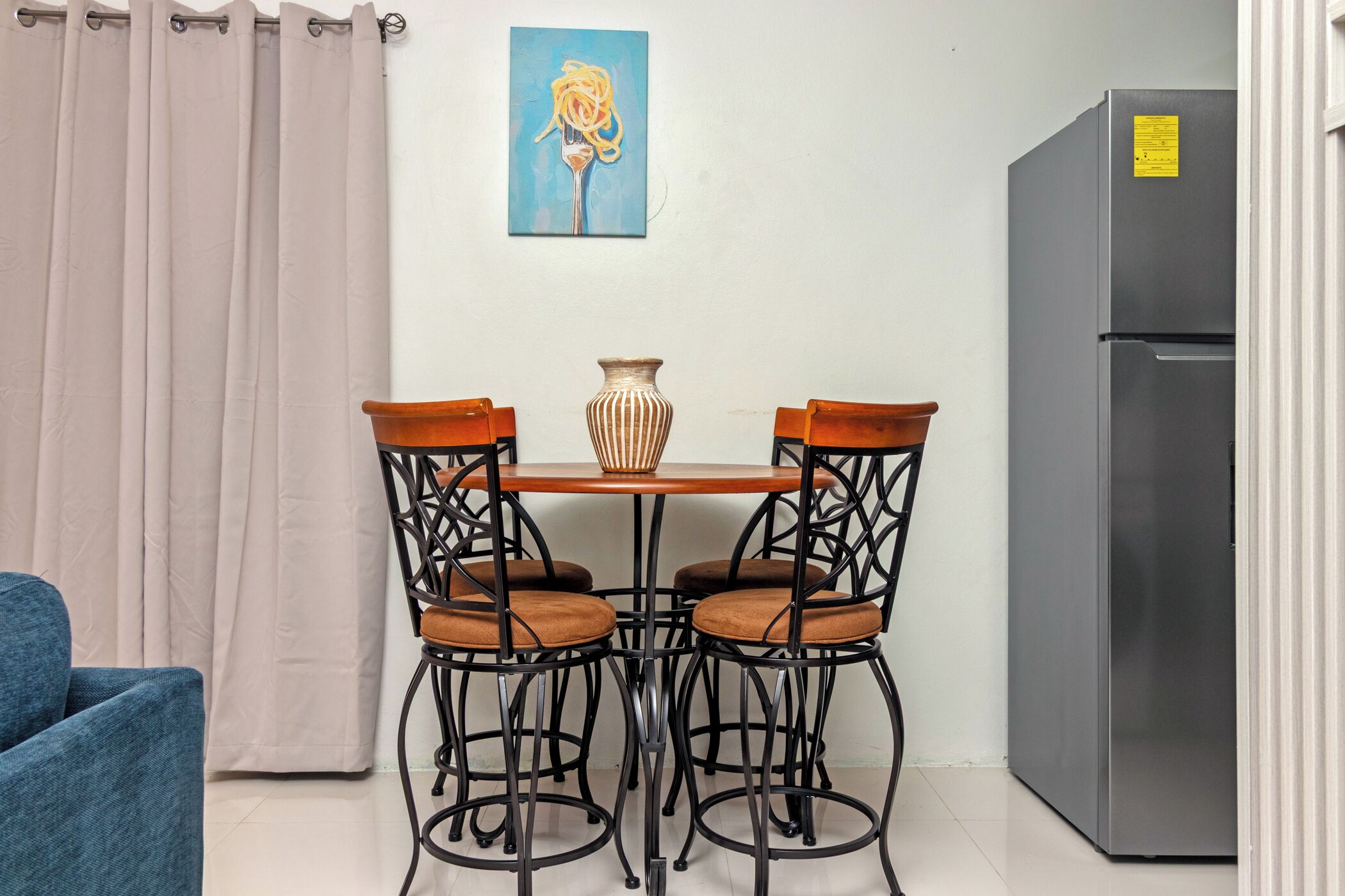 A small kitchenette with a refrigerator, a dining table with four chairs, and a painting on the wall.