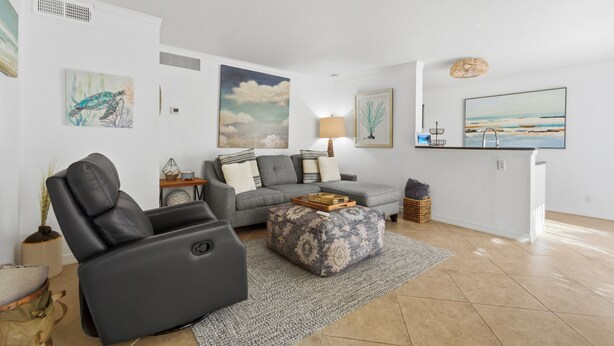 A modern living room with a gray sofa, a black recliner, and a patterned ottoman. There are wall art pieces, a lamp, and a small side table with decorative items.