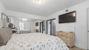 A bedroom with a large bed, a dresser, a TV mounted on the wall, and a sliding glass door leading to a balcony.