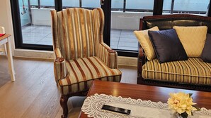 A room with a striped armchair and a sofa with cushions, a wooden coffee table with a remote control and a vase of flowers, and large windows with a view of the street and trees. - Apartment in Windsor (MELBOURNE)