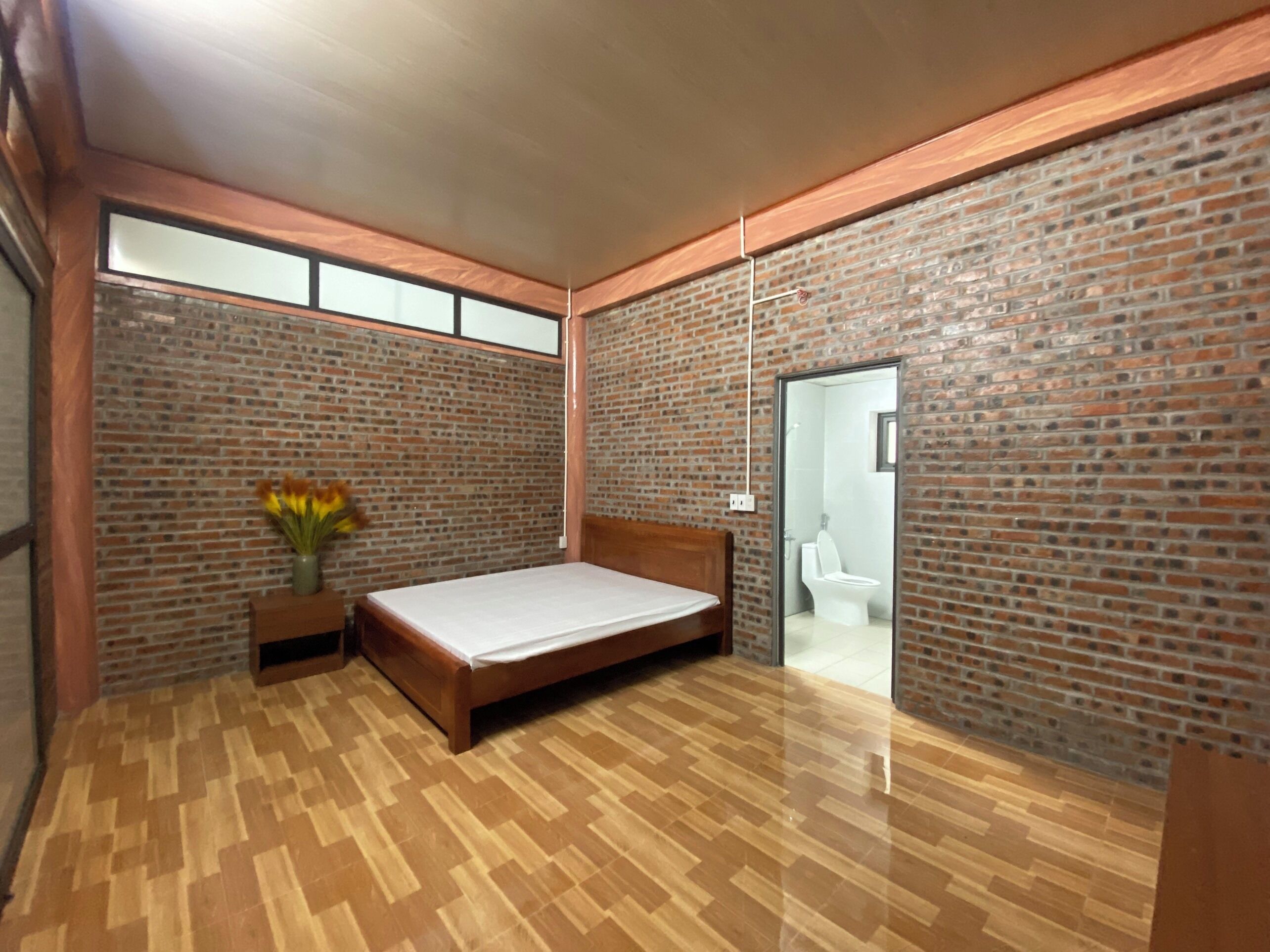 A bedroom with a wooden floor, a bed, a nightstand with a vase of flowers, and brick walls.