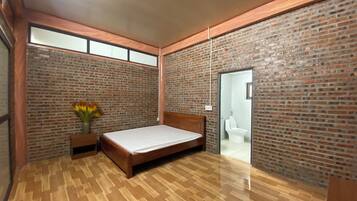 A bedroom with a wooden floor, a bed, a nightstand with a vase of flowers, and brick walls.