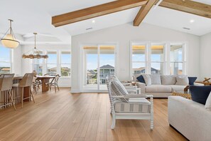 A spacious living room with a wooden floor, a dining area with wicker chairs, a sofa set, and large windows offering a view of the surrounding area. - Fairways & Waves | Ocean Isle Beach, NC (Ocean Isle Beach)