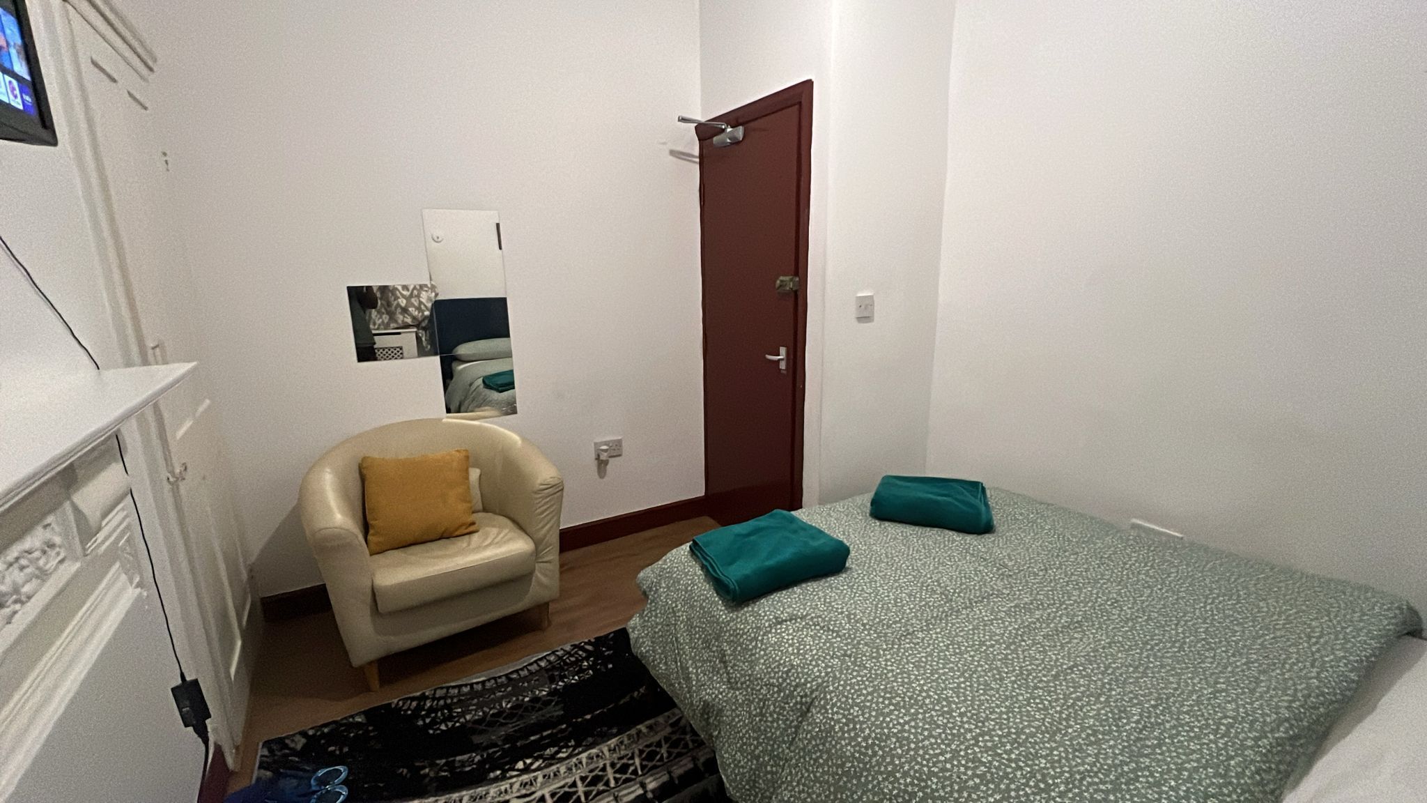 Een kleine, verzorgde slaapkamer met een bed, een stoel, een deur en een spiegel aan de muur.