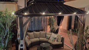 Une terrasse extérieure avec un canapé incurvé, une table basse ronde et une pergola illuminée de guirlandes lumineuses.