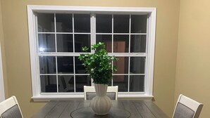 Una mesa de comedor con seis sillas, un jarrón con una planta y una ventana con vistas a una casa.