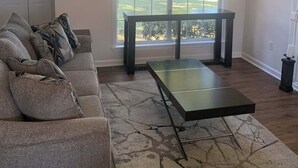 Una sala de estar con sofá, una mesa de centro, un ventilador de techo y un ventanal con vistas al exterior.