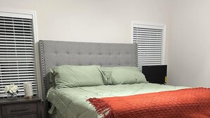 Un dormitorio con una cama tapizada en gris, un edredón rojo y un cabecero también en gris. Se ve un mesita de noche de madera con cajones y una pequeña mesa con un florero y una vela.