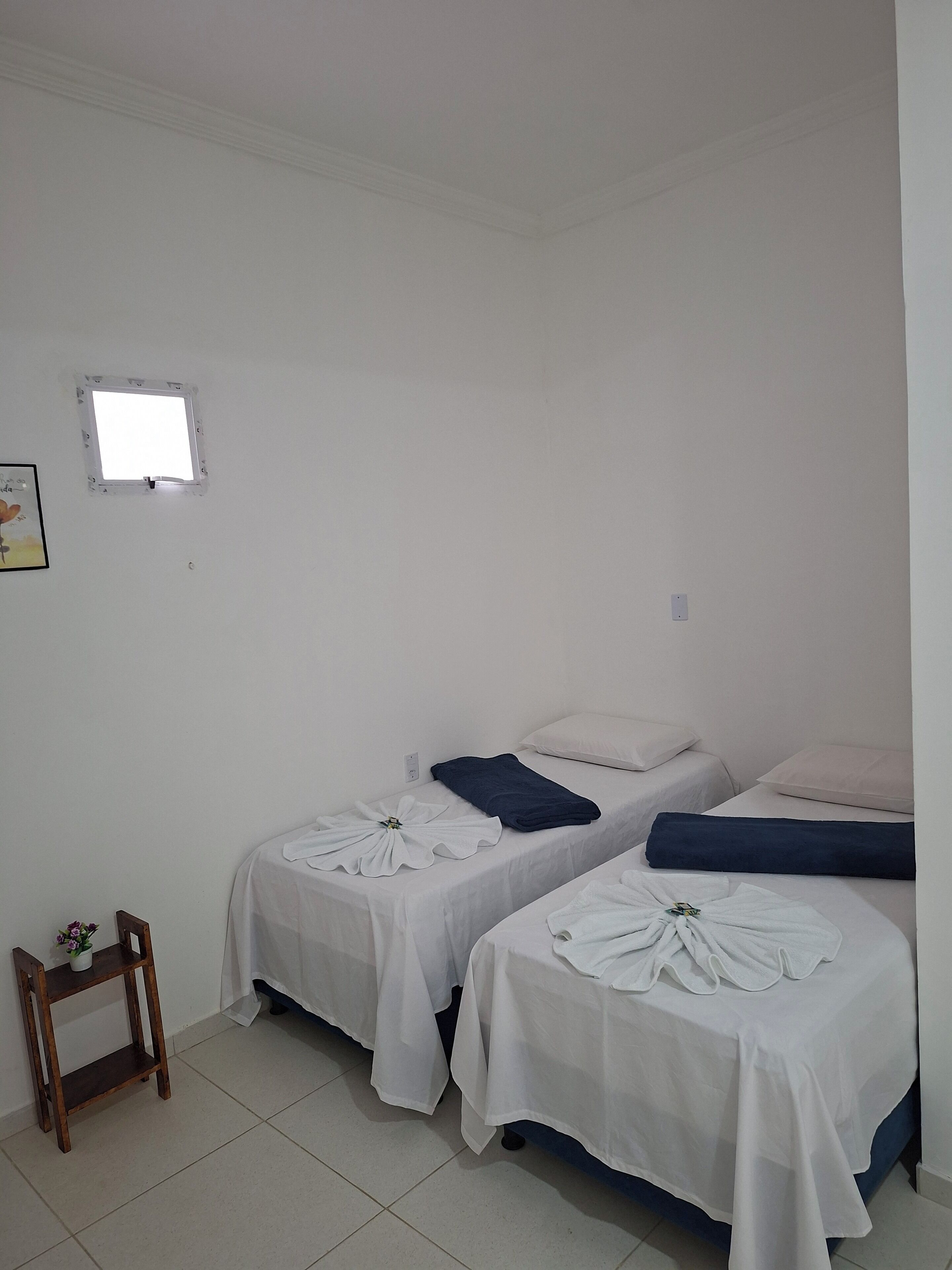 Ein Hotelzimmer mit zwei Betten, jedes mit weißer Bettdecke und blauem Kopfkissen. Über einem Bett befindet sich ein kleines Fenster, ein Holzstuhl und ein Tisch mit einer Topfpflanze.
