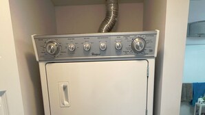 A compact, white, stacked washer and dryer unit in a small laundry room with a door on the left and a sink with toiletries on the right. - Spacious Downtown 1 bedroom condo with Gym, Walk to Old Port (Montréal)