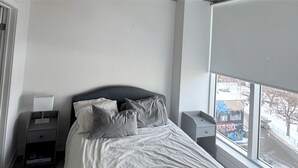 A bedroom with a bed, a nightstand, a drawer, a window with a view of the street, and a door. - Spacious Downtown 1 bedroom condo with Gym, Walk to Old Port (Montréal)