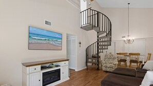 Een woonkamer met een houten vloer, een bruine bank, een wit tv-meubel, een spiltrap en een schilderij met een strandthema.