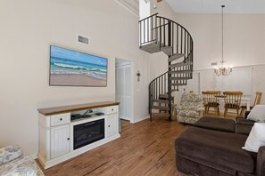 Een woonkamer met een houten vloer, een bruine bank, een wit tv-meubel, een spiltrap en een schilderij met een strandthema.