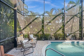 A poolside area with wicker chairs and a table, surrounded by glass walls with a view of palm trees. - Aurora Breeze Estate - The Heart of Matlacha Living (Matlacha)