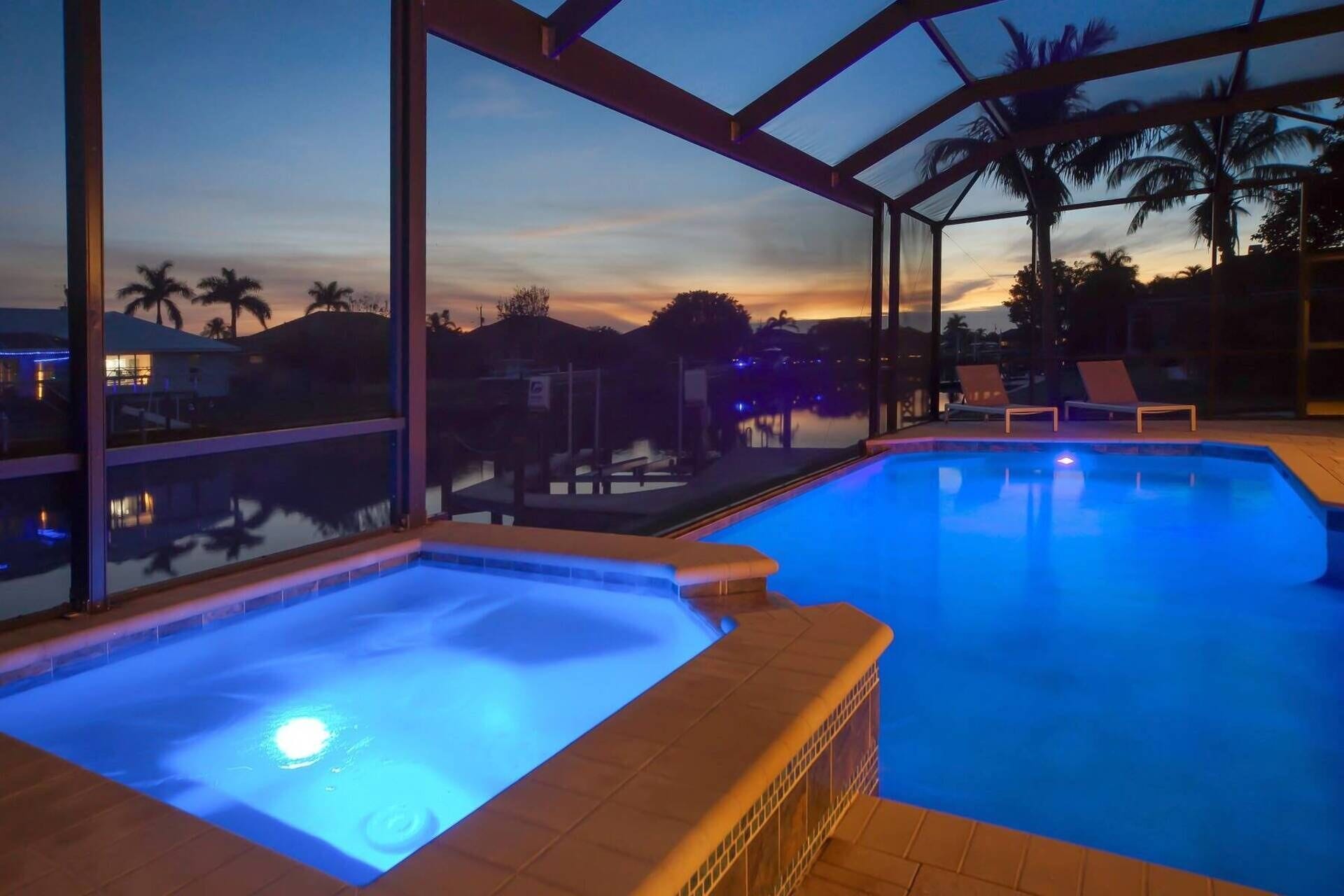A pool area with two hot tubs and lounge chairs, surrounded by a glass enclosure with a view of palm trees and a sunset.