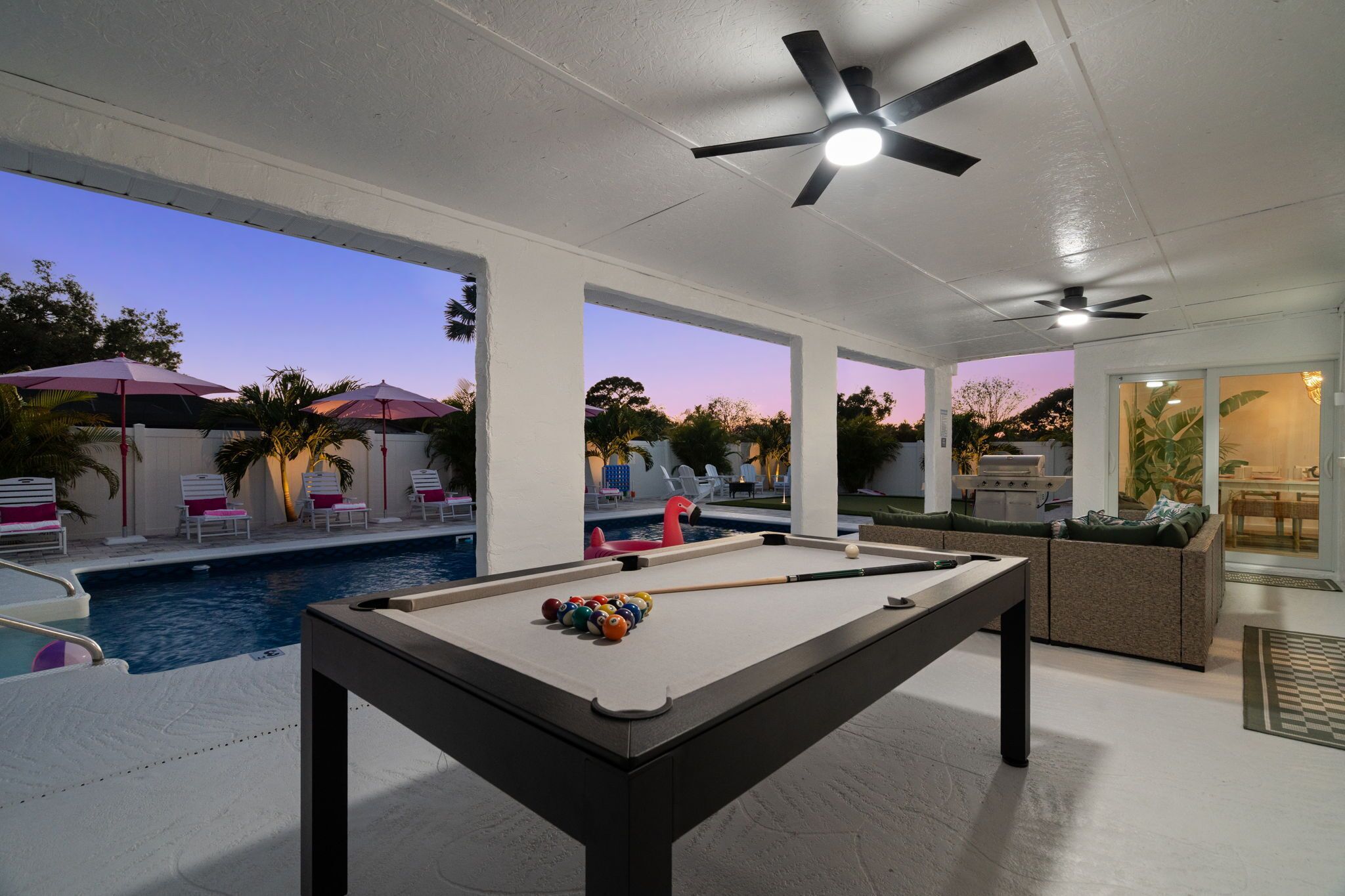 A pool table with balls, a ceiling fan, and a pool in a well-lit indoor space with a view of the sky.