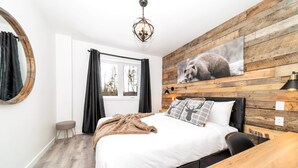 Een slaapkamer met een bed, een houten hoofdbord, een raam met gordijnen, een ronde spiegel, een houten tafel en een wandlamp.