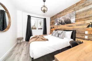 Ein Schlafzimmer mit einem Bett, einem Holzkopfstück, einem Fenster mit Vorhängen, einem runden Spiegel, einem Holztisch und einer wandmontierten Leuchte.