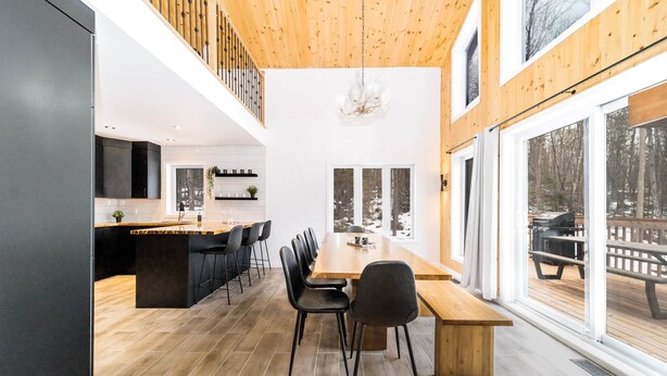 Een moderne keuken met een houten plafond, een eethoek met een lange tafel en stoelen, en grote ramen met uitzicht op sneeuw.