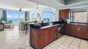 A spacious living area with a large window overlooking the ocean, a kitchen with wooden cabinets, and a dining area with a table and chairs. - Majestic Sun B614*Gulf Views*Heated Pools/Hot Tubs*Beach Gear*Gym*Pickleball (Miramar Beach)