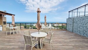 A rooftop patio with round tables and wicker chairs, overlooking a cityscape and the sea. - COSTE0110 - Concept Life Stella (Salvador)