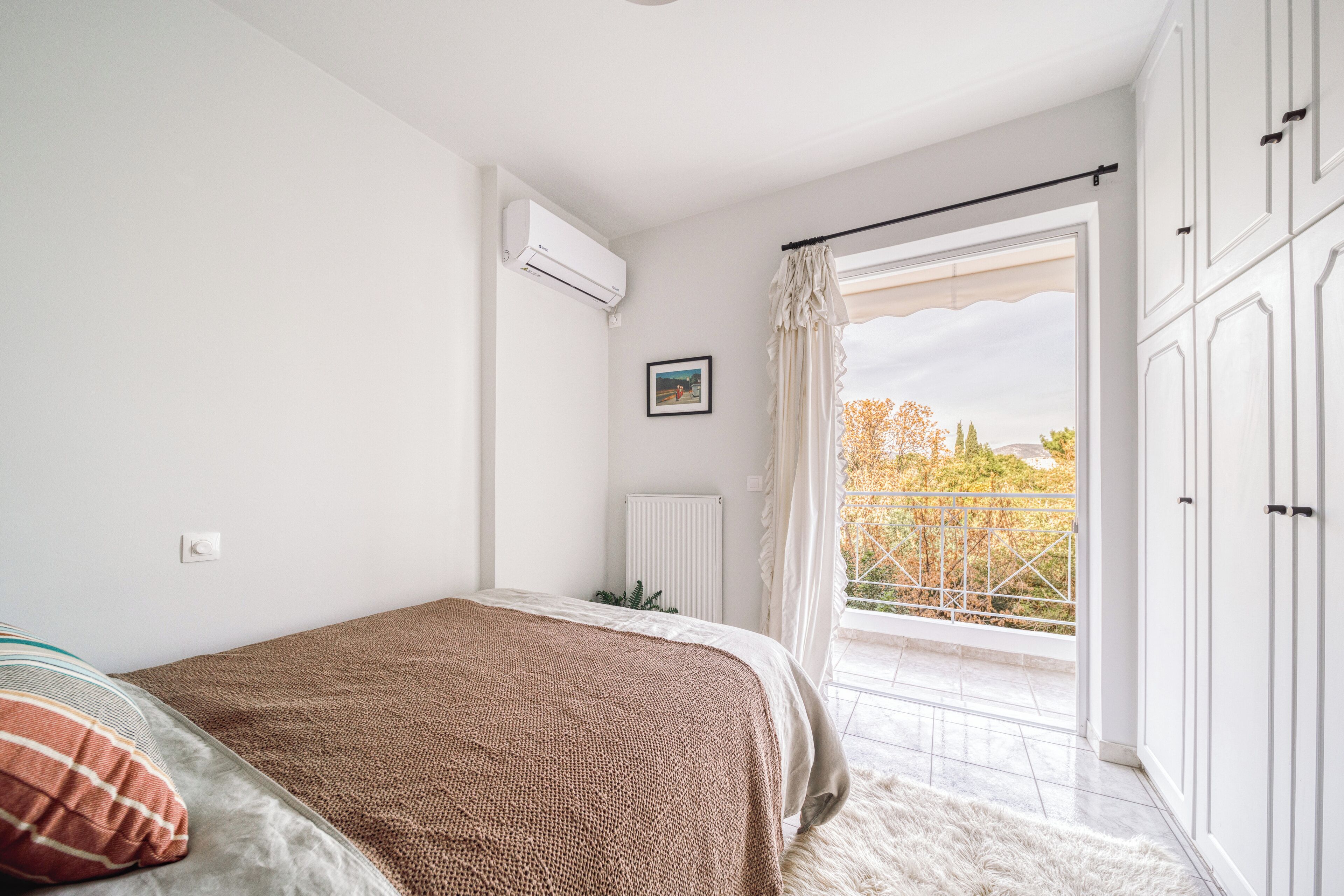 Een slaapkamer met een bed, een radiator, een raam met gordijnen en uitzicht op bomen.