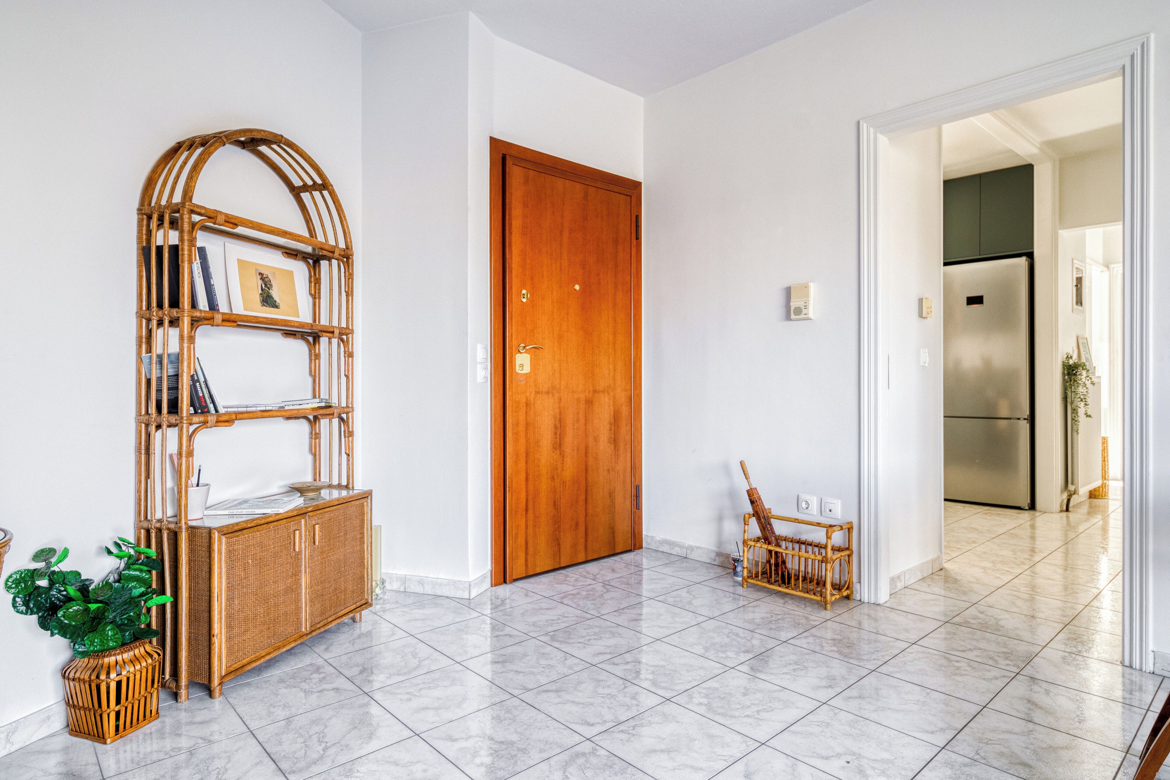 Un pasillo con una puerta de madera, un estante de mimbre y un suelo de baldosas.