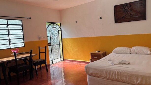 A hotel room with a bed, a desk with chairs, a window with bars, and a door with a decorative glass panel.