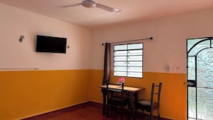 A room with a ceiling fan, a TV mounted on the wall, a window with curtains, a table with chairs, and a door with a decorative glass panel.
