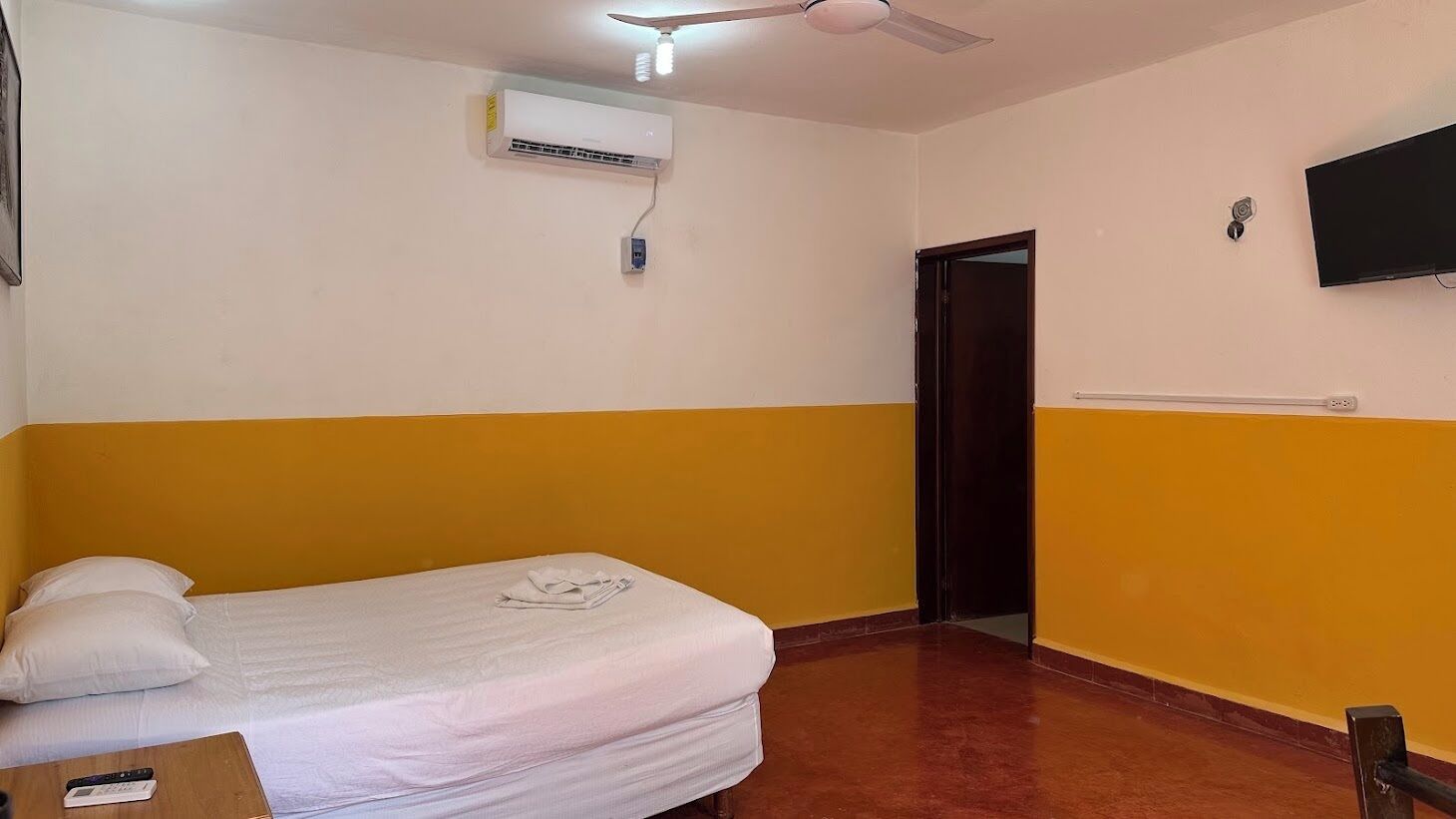 A room with a bed, a ceiling fan, an air conditioning unit, a television mounted on the wall, and a door.