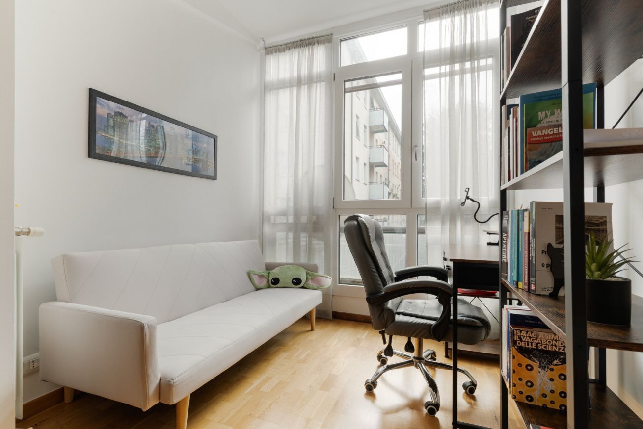 Ein modernes Zimmer mit einer weißen Couch, einem schwarzen Bürostuhl, einem Bücherregal und einem Fenster mit durchsichtigen Vorhängen.