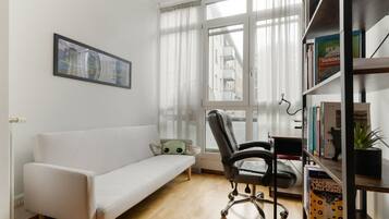 Ein modernes Zimmer mit einer weißen Couch, einem schwarzen Bürostuhl, einem Bücherregal und einem Fenster mit durchsichtigen Vorhängen.