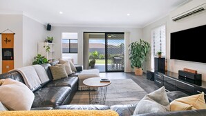 A modern living room with a leather sofa, a coffee table, and a TV. There is a sliding glass door leading to a balcony with plants. - Dakabin Escape Modern Comfort near Dining and Shops (Dakabin)