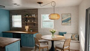 A kitchen with teal cabinets, a wooden countertop, a round dining table with chairs, a hanging light fixture, and a window with blinds.
