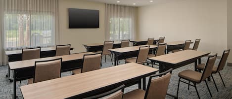 A conference room with multiple tables and chairs, large windows with curtains, and a flat-screen TV mounted on the wall.