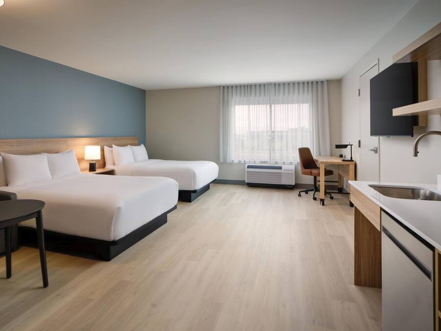 A hotel room with two beds, a desk, a chair, a small table, a TV, and a kitchen area.