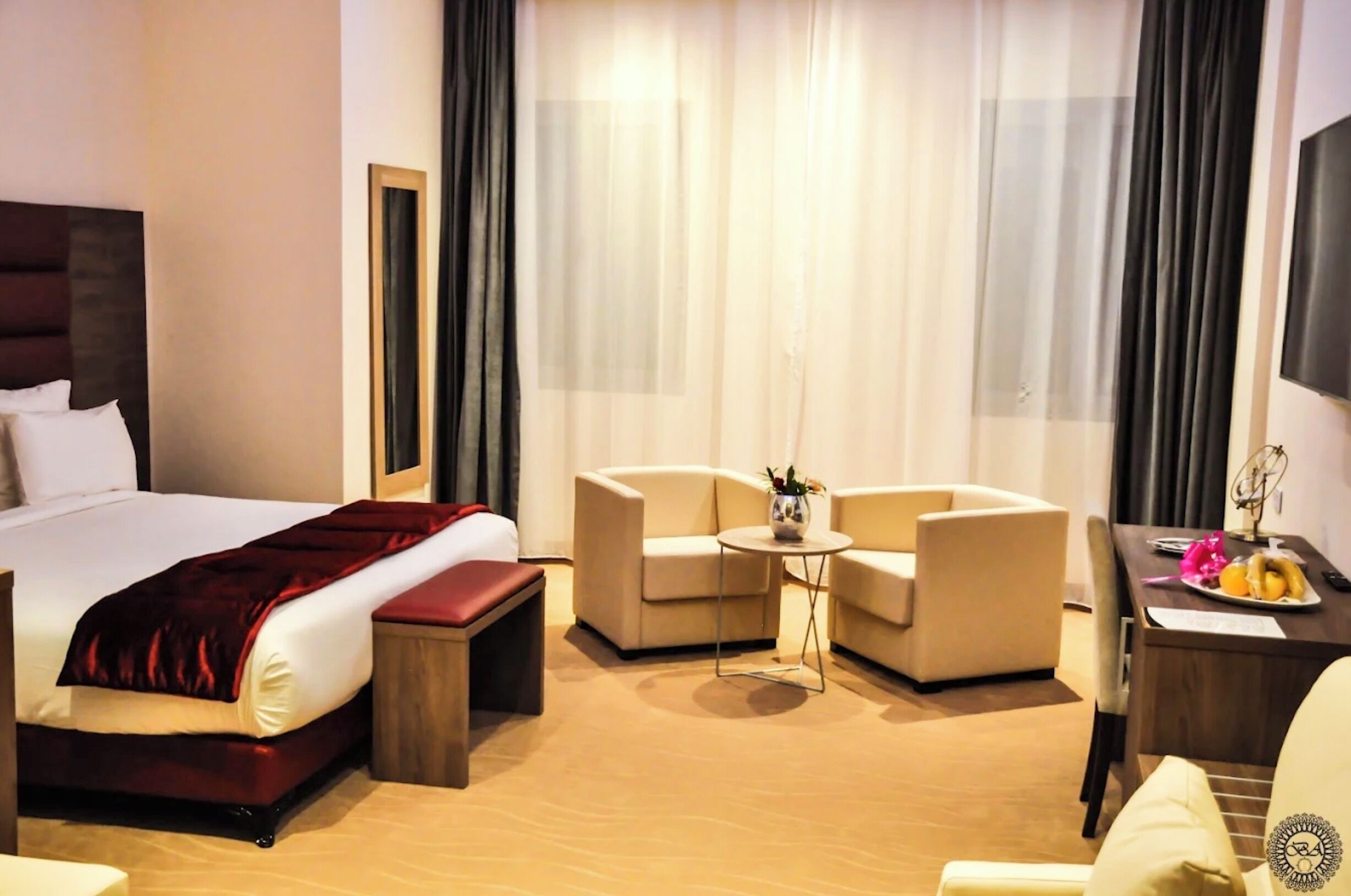 A hotel room with a large bed, two armchairs, a small table with a vase, and a dining table with fruit.