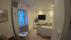 A compact room with a bed, a small table, a window with blinds, a kitchenette with a TV, and a radiator. - Studio "A deux pas des thermes" in the heart of Uriage park. (Saint-Martin-d'Uriage)