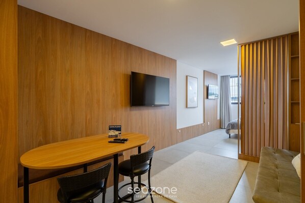 Quarto de hotel moderno com parede de madeira, televisão de tela plana, área de jantar com mesa e cadeiras, e um sofá.