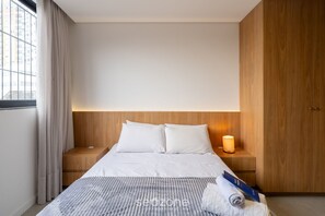 Quarto de hotel com uma cama grande, cabeceira de madeira, mesa de cabeceira com abajur e vista da cidade pela janela.