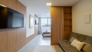 Et moderne hotelværelse med et fladskærms-tv på væggen, en sofa med pude og et stort vindue med lodrette persienner.