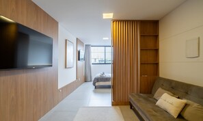 Quarto de hotel moderno com televisão de tela plana na parede, um sofá com almofada e uma grande janela com persianas verticais.