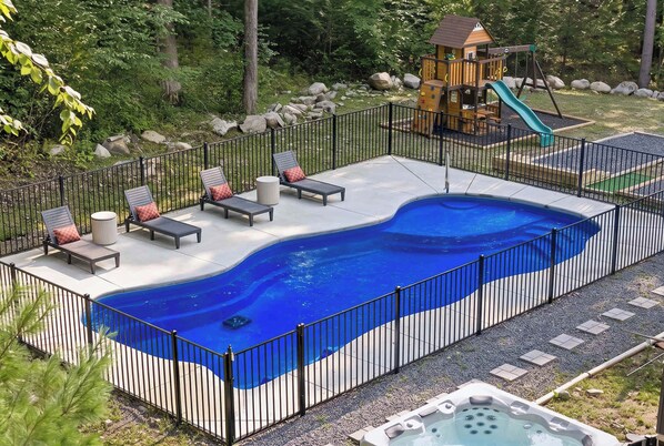 Una piscina con un jacuzzi, rodeada por una valla, y una estructura de juegos infantiles al fondo.