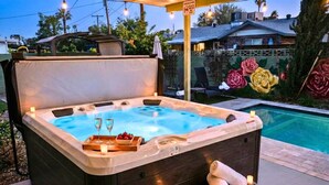 Un bain à remous avec un banc en bois, un plateau avec des verres et des fruits, et une piscine en arrière-plan.