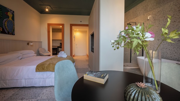 A hotel room with a bed, a chair, a table with a book and a vase with flowers, and a wall with a painting. - Charming elegant room in central Acireale with serene garden view (Acireale)
