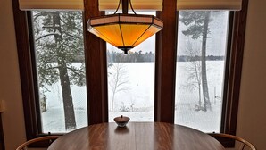 A round wooden dining table with a bowl in the center, flanked by two chairs, set against a window with a view of a snowy landscape and trees. - Foster's Lakeside - Sandy Shore with Snowmobile Access & Year-Round Fun (Tomahawk)